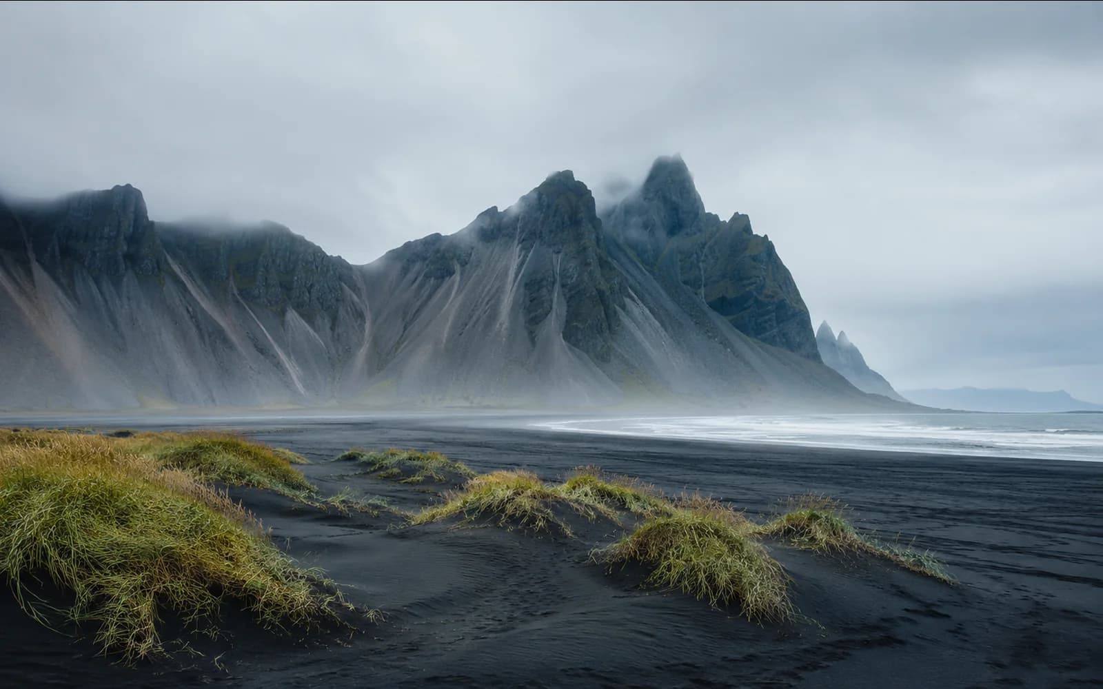 Icelandic black-sand coastal landscape with misty jagged mountains and grassy dunes